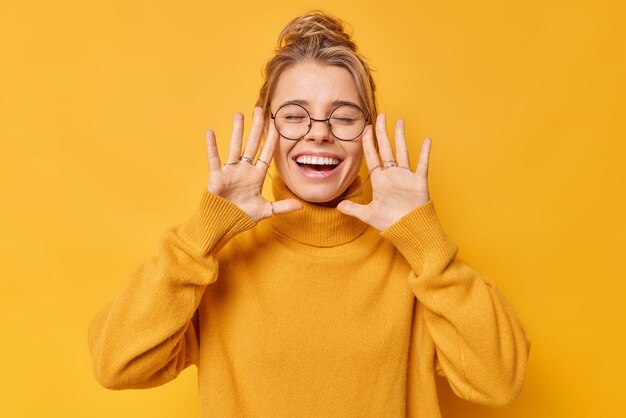 Froh positive junge Frau hält Handflächen in der Nähe von Gesicht erhoben Ausrufe glücklich schließt die Augen trägt lässige Pullover lächelt breit zeigt weiße Zähne isoliert über gelbem Hintergrund Konzept der menschlichen Emotionen