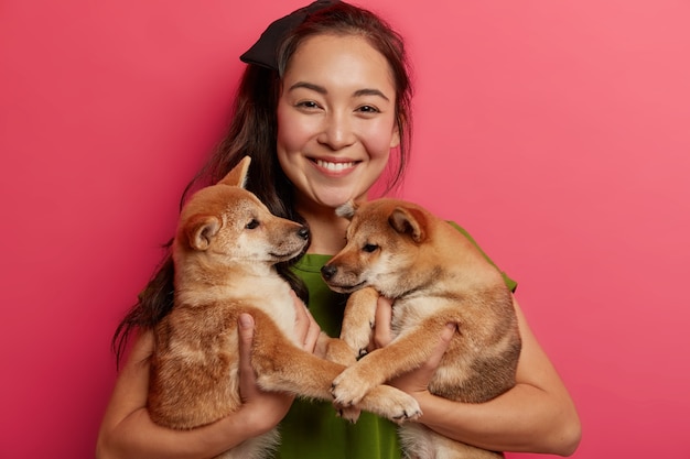 Frohe asiatische Frau posiert mit zwei kleinen Welpen, mag Shiba Inu Hunde, lächelt breit, bekommt gute Nachrichten vom Tierarzt, glücklich, gesunde Haustiere zu haben.