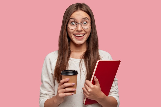 Frohe brünette frau mit aufgeregtem ausdruck, sieht glücklich aus, trägt eine runde brille, hält ein rotes lehrbuch und nimmt kaffee heraus, reagiert auf gute nachrichten von klassenkameradin, isoliert auf rosa wand