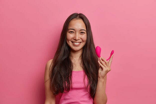 Frohe brünette Frau posiert mit intelligentem Bluetooth-Vibrator, verwendet spezielle App auf dem Handy, um den Orgasmus zu verbessern, hält Sex-Tool, um das Vergnügen zu verbessern, isoliert auf rosa Wand. Moderne Technologien und Sexualleben