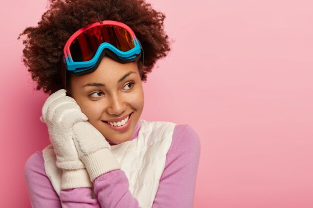 Frohe dunkelhäutige Frau mit Afro-Frisur, trägt eine Snowboardbrille, weiße weiche Handschuhe, genießt Wintersport, schaut glücklich weg, isoliert über rosa Hintergrund, fühlt sich verspielt und entzückt