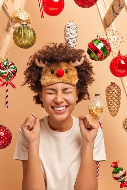 Frohe dunkelhäutige Frau mit lockigem buschigem Haar hebt geballte Fäuste und feiert Erfolg bereitet sich auf das Feiern von Weihnachten vor trägt Freizeitkleidung Posen