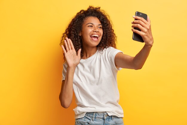 Frohe dunkelhäutige glückliche Frau mit Afro-Frisur, hält modernes Handy vor Gesicht, Wellen Handfläche in der Kamera, macht Videoanruf, gekleidet in Freizeitkleidung