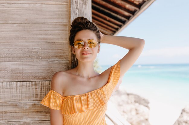 Frohe europäische Frau trägt elegante Brille, die im Resort chillt. Erfreutes braunhaariges Mädchen in orangefarbener Kleidung, das im Strand aufwirft