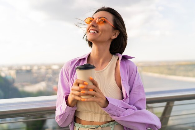 Frohe Frau mit kurzen Haaren im stilvollen Sommeroutfit trinkt Kaffee auf der modernen Brücke