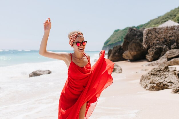Frohe Frau trägt Band und Sonnenbrille, die am wilden Strand tanzen. Foto im Freien des erfreuten gebräunten Mädchens, das positive Gefühle im Strand ausdrückt
