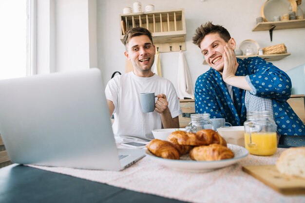 Frohe Freunde, die Frühstück beim Aufpassen etwas auf Laptop genießen