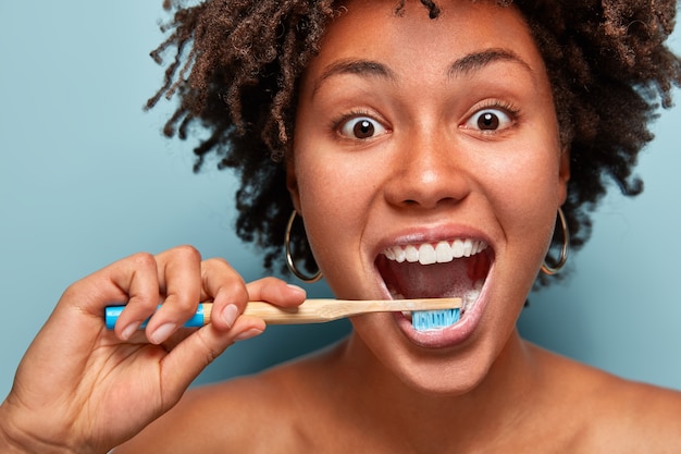 Frohe fröhliche Frau mit dunkler, gesunder Haut, zufrieden mit neuer Zahnpasta, reinigt die Zähne, öffnet den Mund weit, hat einen glücklichen Ausdruck, lockiges Haar, Modelle über der blauen Wand. Nahaufnahme Porträt.