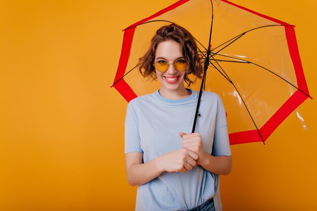 Frohe junge Frau im blauen T-Shirt, das stilvollen Sonnenschirm hält. Innenporträt des erfreuten lockigen Mädchens in der Sonnenbrille, die mit Lächeln unter Regenschirm aufwirft.