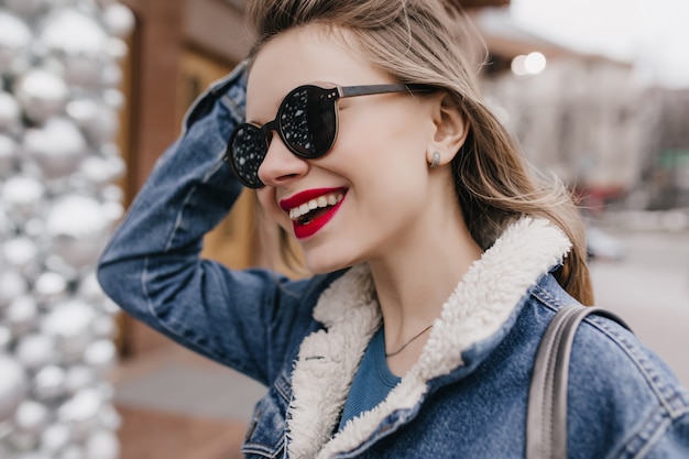 Kostenloses Foto frohe junge frau in jeanskleidung, die ihre haare berührt, während sie durch die stadt geht. außenaufnahme des schönen mädchens in der guten stimmung, die im frühlingstag entspannt.