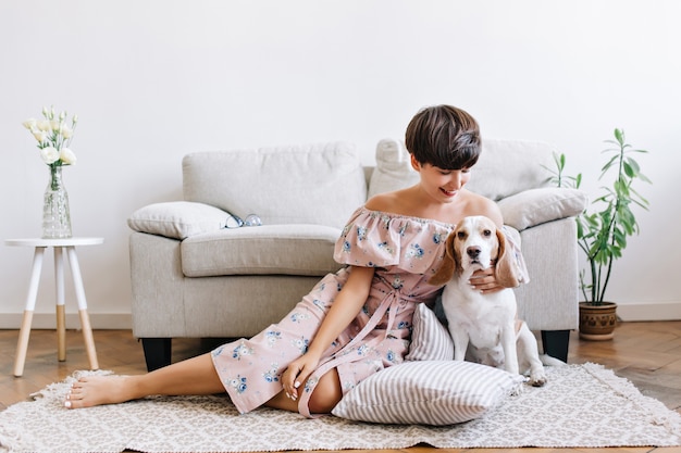 Frohe junge Frau mit glänzendem braunem Haar, das mit ihrem niedlichen Beagle-Welpen auf dem Boden aufwirft. Innenporträt des aufgeregten Mädchens im Kleid mit Blumendruck, der auf dem Teppich mit Hund sitzt