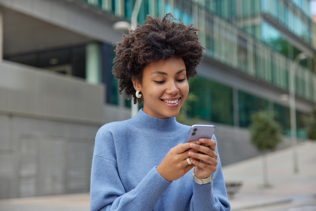 Frohe junge Frau mit lockigem, buschigem Haar hält moderne Handy-Downloads erstaunliche Anwendung surft in sozialen Netzwerken trägt einen lässigen blauen Pullover