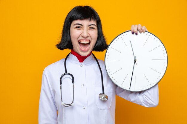 Frohe junge hübsche kaukasische Frau in Arztuniform mit Stethoskop, die Uhr hält