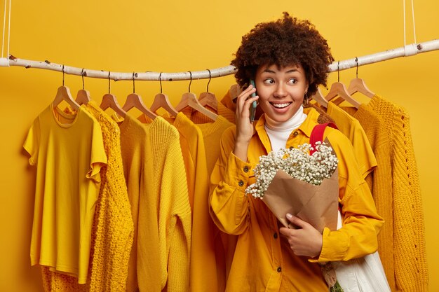 Frohe lockige Frau mit fröhlichem Gesichtsausdruck, ruft Freund an, hält schönen Blumenstrauß, trägt Tasche, posiert gegen gelbe helle Kleidung auf Lumpen