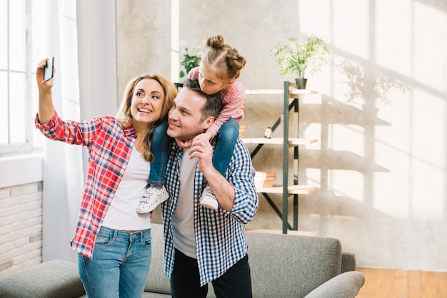 Frohe Mutter, die selfie am Handy während Vater trägt, der ihre Tochter auf Schulter im Wohnzimmer trägt