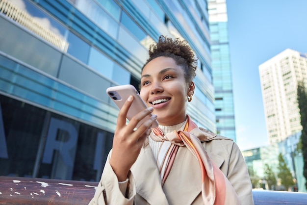 Frohe schöne junge Frau in Mantel und Kopftuch um den Hals, die Sprachanrufe aufzeichnet, hält das Smartphone in der Nähe des Mundes vor dem Hintergrund der modernen Stadt Moderne Technologien und Lifestyle-Konzept