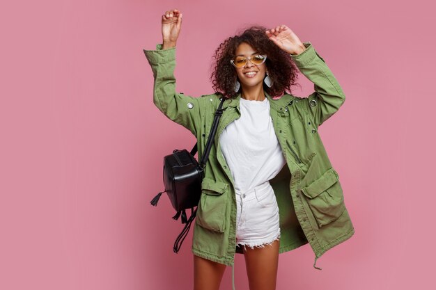 Frohe schwarze Frau, die Spaß über rosa Wand hat. Weißes T-Shirt, grüne Jacke. Stilvoller Frühlingslook.