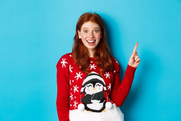 Frohe Weihnachten. Fröhliches rothaariges Mädchen im Weihnachtspullover, das mit dem Finger auf die obere rechte Ecke zeigt und aufgeregt lächelt und Neujahrspromo zeigt.