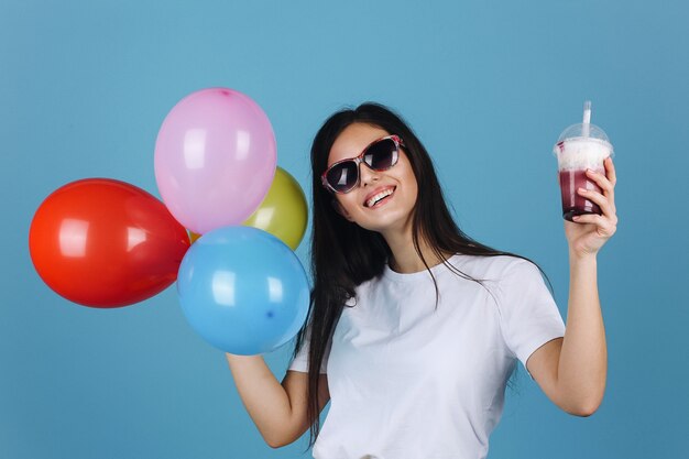 Froher Brunette in der schwarzen Sonnenbrille schaut die glückliche Aufstellung mit einem Cocktail und Ballonen