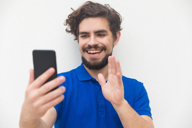 Froher freundlicher Kerl mit dem Smartphone, der hallo wellenartig bewegt