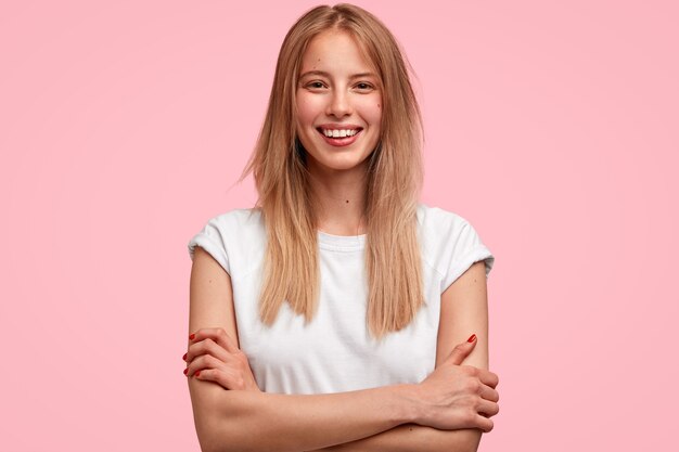 Frohes blondes Frauenmodell hält Arme verschränkt, hat ein zahniges Lächeln, freut sich über das Treffen mit dem besten Freund, teilt Eindrücke nach der Party, gekleidet in weißes T-Shirt