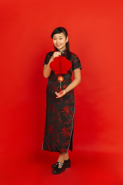 Frohes chinesisches Neujahr. Asiatisches junges Mädchenporträt lokalisiert auf rotem Hintergrund. Weibliches Modell in traditioneller Kleidung sieht glücklich aus und lächelt mit Dekoration. Feier, Urlaub, Emotionen.