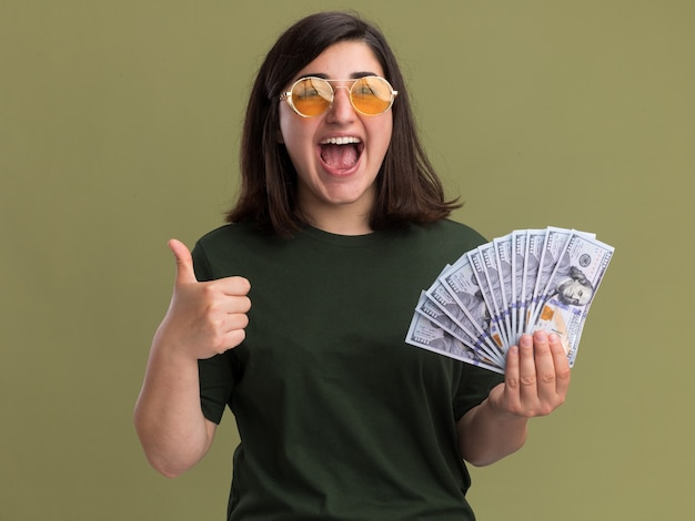 Frohes junges hübsches kaukasisches Mädchen in Sonnenbrillen Daumen hoch und hält Geld