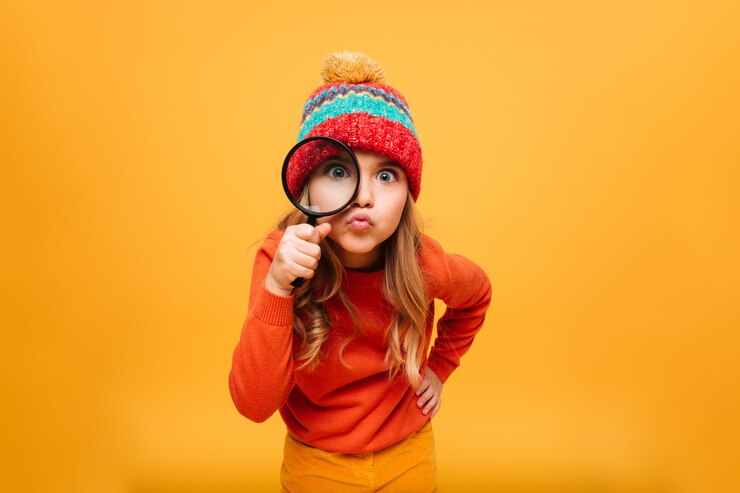 Kostenlose Foto frohes junges mädchen in der strickjacke und in hut, welche die kamera mit vergrößerungsglas über orange betrachten