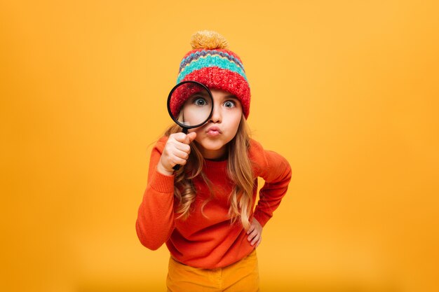 Frohes junges Mädchen in der Strickjacke und in Hut, welche die Kamera mit Vergrößerungsglas über Orange betrachten