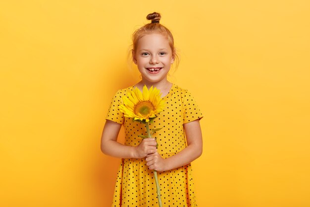 Frohes kleines rothaariges Mädchen, das mit Sonnenblume im gelben Kleid aufwirft