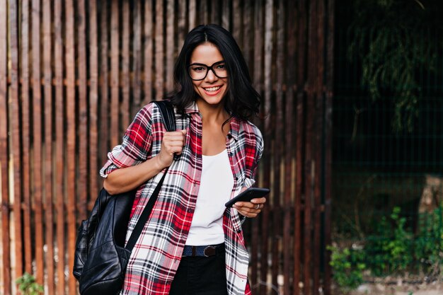 Frohes lateinisches weibliches Modell, das positive Emotionen während des Fotoshootings auf Holzwand ausdrückt. Entspanntes brünettes Mädchen, das Rucksack und Telefon hält.