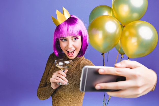 Frohes neues Jahr Feier Momente der aufgeregten jungen Frau mit rosa Haarschnitt machen Selfie Porträt. Luxuskleid, goldene Luftballons, Alkoholcocktail, Geburtstagsfeier.