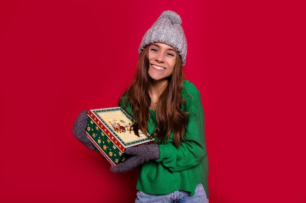 Frohes neues Jahr Partyzeit der lächelnden schönen jungen Frau, die ein Geschenk zur Kamera auf rotem Hintergrund hält. Nettes Lächeln, Winterpullover und Mütze, Spaß haben, Geburtstagsfeier