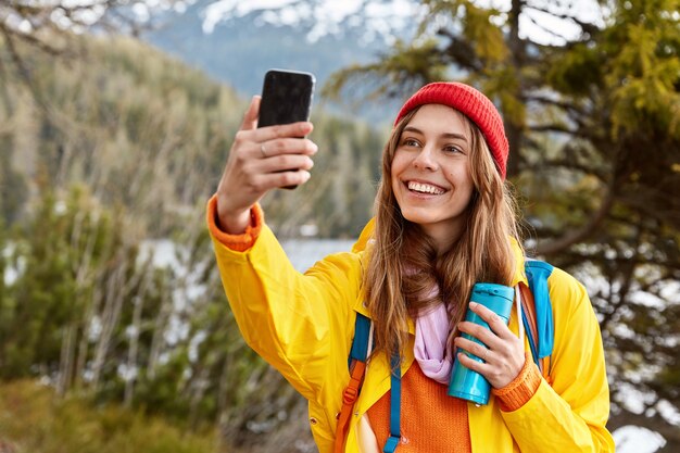 Frohes schönes dunkelhaariges Mädchen macht Selfie-Porträt auf Handy, gekleidet in Regenmantel, Kopfbedeckung