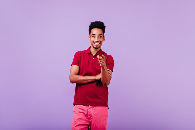 Frohes schwarzes Mann im Sommeroutfit, das auf violetter Wand aufwirft. sorgloser brünetter Kerl, der gute Gefühle ausdrückt.