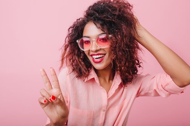 Frohes sinnliches Mulattenmädchen in der stilvollen Sonnenbrille, die mit Friedenszeichen aufwirft. Elegante charmante Frau in formeller Kleidung, die Spaß hat.