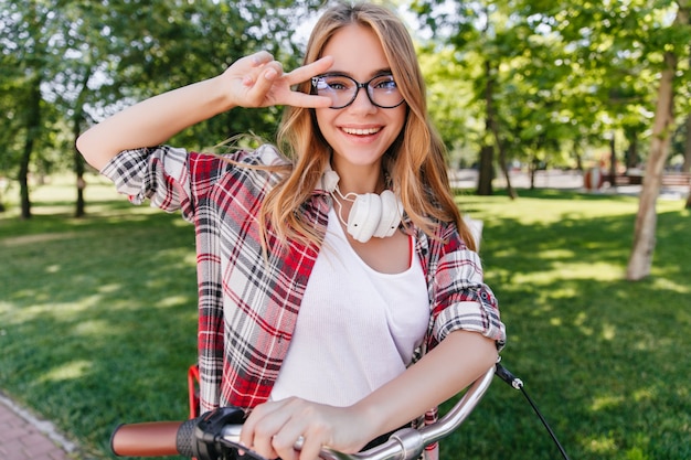 Frühlingsfoto des niedlichen kaukasischen Mädchens mit dem Fahrrad. Außenaufnahme des weiblichen Debonair-Modells in Brille und in den Kopfhörern.