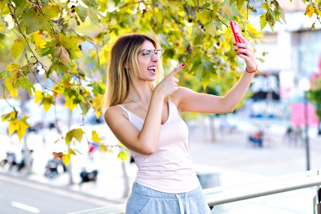 Frühlingslebensstilporträt der hübschen blonden Frau, die selfie macht und auf Video-Chat mit ihrem Freund, lässige sportliche Kleidung, sonnige Pastellfarben spricht.