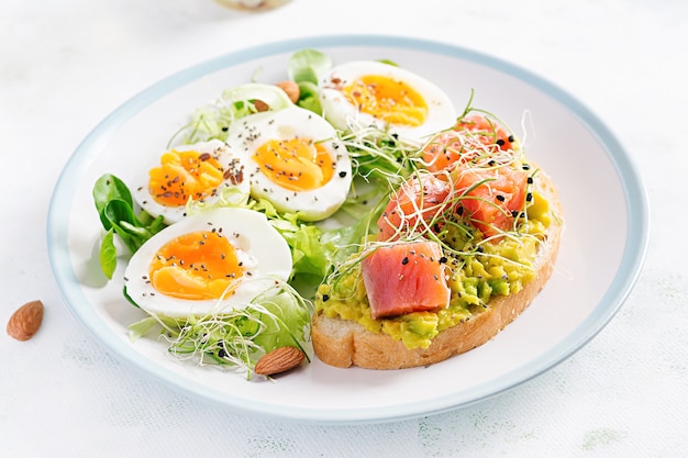 Frühstück. Gesundes offenes Sandwich auf Toast mit Avocado und Lachs, gekochten Eiern, Kräutern, Chiasamen auf weißem Teller mit Kopierraum. Gesunde Eiweißnahrung.