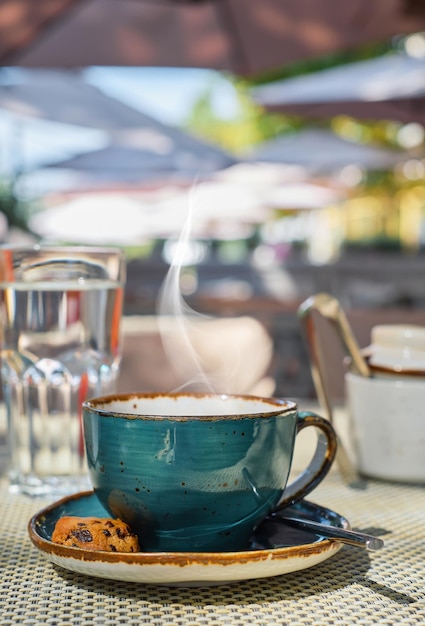 Frühstück in einem Café vertikaler Schussdampf steigt über einer Tasse Espressokaffee, Glas Wasser und Keksen auf dem Tisch, Nahaufnahme, Sonnenlicht und selektiver Fokus, leere Raumidee für Werbung oder Banner