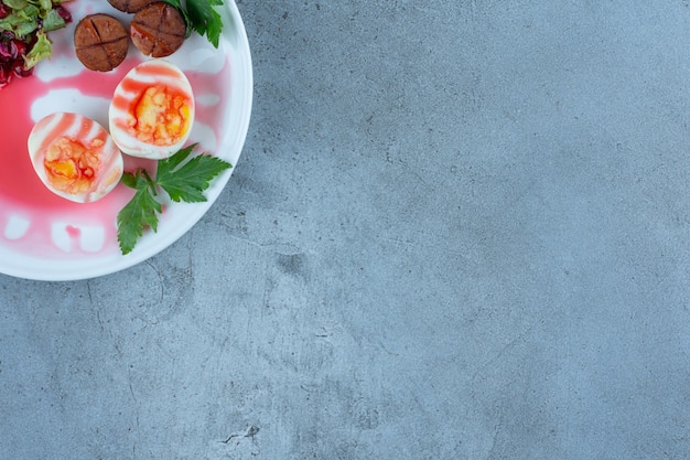 Frühstücksplatte mit gekochten Eiern, gebratenen Wurstscheiben und einer kleinen Portion Granatapfelsalat auf Marmor.