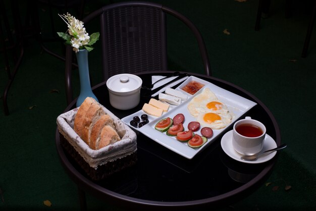 Frühstückstablett mit Spiegeleiern, Würstchen, Käse, Marmelade, Butter, Brot und einer Tasse Tee.