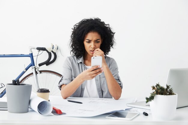 Frustrierte junge dunkelhäutige Frau, die am weißen Tisch sitzt und neues Projekt hält, das Handy hält