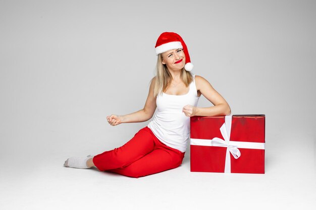 Frustrierte junge Frau in Sankt-Hut, die ihre Fäuste ballt, während sie neben rot verpacktem Weihnachtsgeschenk sitzt. Platz kopieren