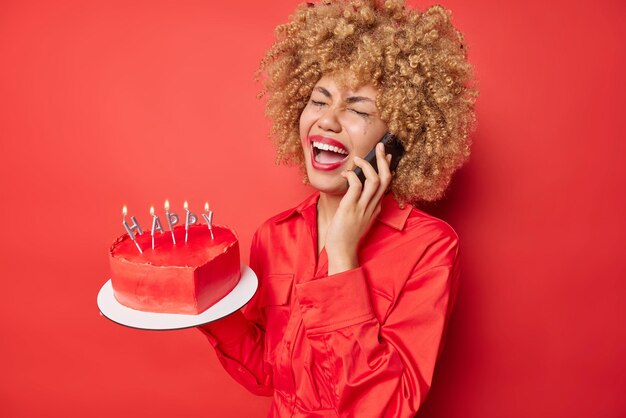 Frustrierte junge lockige Frau hat Telefongespräche aus Verzweiflung trägt Hemd hält Herzkuchen mit brennenden Kerzen umgekippt, um Geburtstag allein isoliert über rotem Hintergrund zu feiern