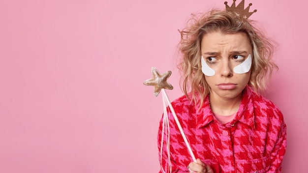 Kostenloses Foto frustrierte, unglückliche frau sieht traurig weg, hat schlechte laune, nachdem sie geweint hat, verwöhntes make-up trägt schönheitsflecken auf, trägt eine kleine krone und eine karierte jacke hält zauberstab-posen an der rosa studiowand