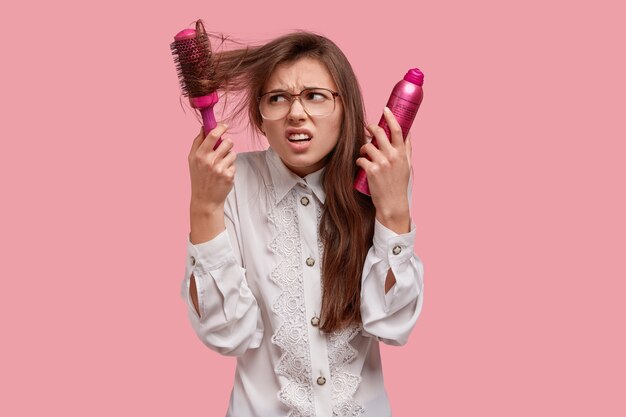 Frustrierte Unzufriedenheit Frau versucht, Haarbürste aus den Haaren zu blasen, hält Haarspray, hat Probleme mit Haaren, gekleidet in weißes Hemd Stirnrunzeln Gesicht mit Missfallen auf rosa Wand isoliert. Schönheitskonzept