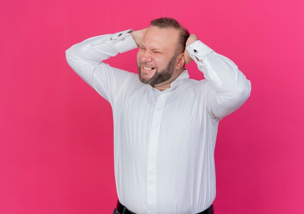 Frustrierter bärtiger Mann, der weißes Hemd trägt, das seinen Kopf berührt, der wild über rosa Wand steht