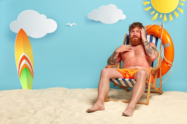 Frustrierter bärtiger rothaariger Mann bekommt am Strand einen Sonnenbrand