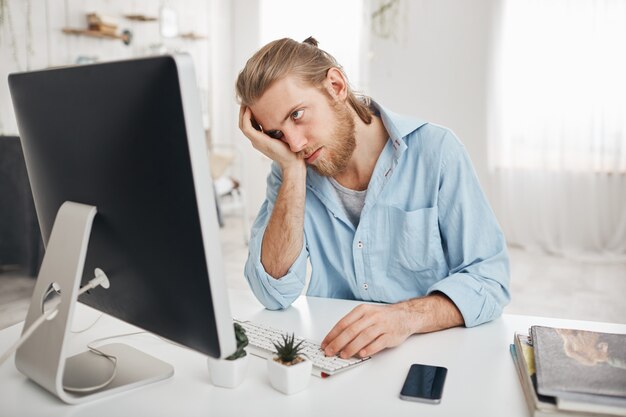 Frustrierter müder bärtiger kaukasischer Angestellter, der seinen Kopf berührt, sich wegen Überlastung absolut erschöpft fühlt, Konten berechnet und vor dem Computerbildschirm sitzt. Frist und Überarbeitung